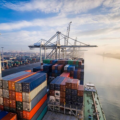 Containerstapel und Kräne im Hafen von Rotterdam an einem Fluss unter leicht bewölktem Himmel.
