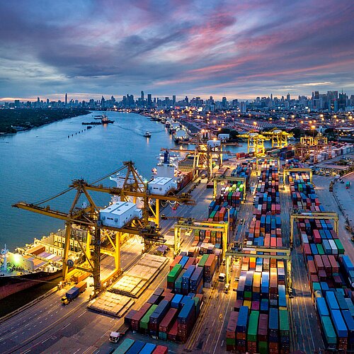 Luftaufnahme Hafen mit Kran, der Container auf Schiff lädt, vor Kulisse nächtlicher Skyline von Bangkok.